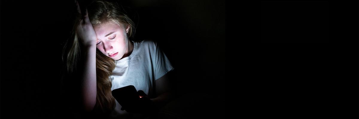 Ung pige sidder i mørkt rum. Hovedet hviler i den ene hånd. Den anden hånd holder en telefon, som lyser på hendes ansigt.