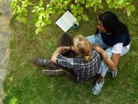 Mand og kvinde sidder på græsset med bog foran sig. Kvinden holder manden på skulderen.