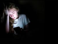 Ung pige sidder i mørkt rum. Hovedet hviler i den ene hånd. Den anden hånd holder en telefon, som lyser på hendes ansigt.